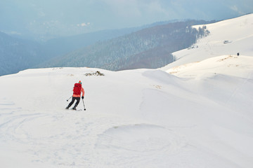 Fototapeta na wymiar Off piste skier, wearing colorful red clothes, descending on a ridge in Baiului mountains, Romania, towards the foggy forest - copy space on the right.