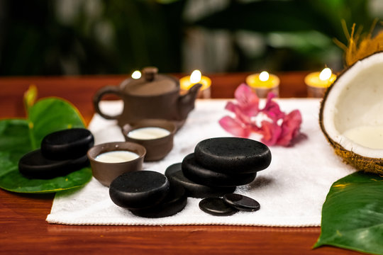 A terry towel on which there is a clay teapot and cups for drinks with milk, stones for stone therapy, lighted candles, a mangolia flower and a twisted terry towel on a wooden table
