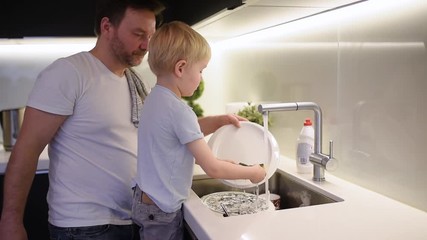 Little boy washing dirty dishes. Child cleans crockery while doing cleaning at home. Dad's little helper. Housework for kids.