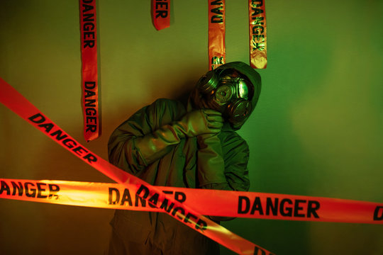 A Guy In A Protective Suit With A Gas Mask On His Face And A Hood On His Head And Long Rubber Protective Gloves Posing Standing Near A Green Wall With Hanging Danger Tapes And Showing Suffocation.