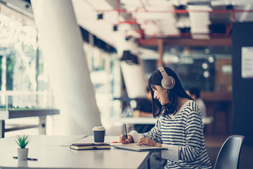 Young Asian girl listening the music with headphones and reading books while sitting at the table in the modern working space.