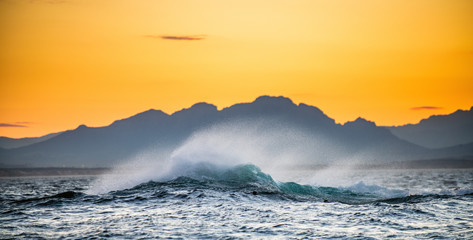Fototapeta na wymiar Seascape. Red dawn sky, waves crashing with splashes against stones, silhouettes of mountains on the horizon. South Africa.