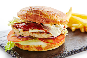 Fresh Chicken Burger with Onions, Tomato Sauce, Pickled Cucumbers, Green Lettuce, Turkey, Cheese and French Fries Garnish. Hamburger or Chickenburger on Natural Black Stone Plate Isolated on White