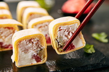 Chicken burrito with smoked fillet, cream cheese, tomato, pickled cucumbers and cheese cake. Macro shot of chopsticks holding chicken maki sushi roll or tortilla rolls on black background © Ryzhkov