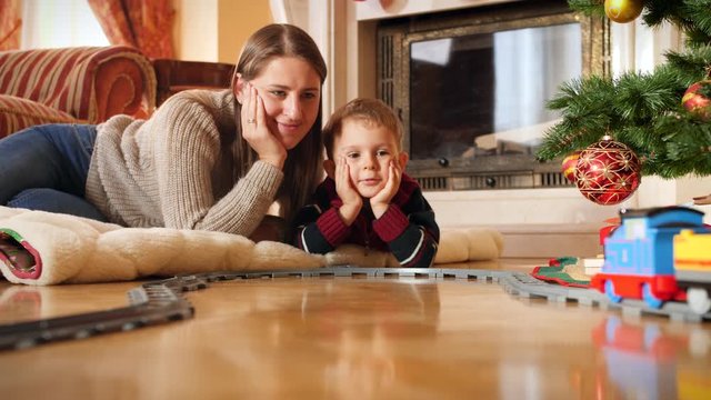 4k video of happy little boy lying with his mother on floor at living room on Christmas morning and looking how his new toy train riding on railroad. Child receiving gifts and presents from Santa