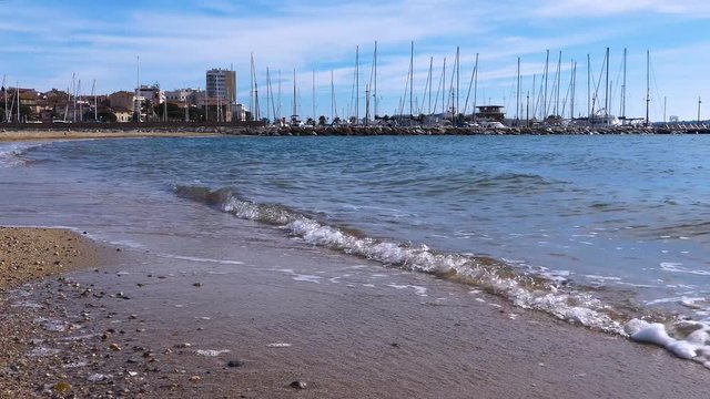 French Riviera. Southern France. Mediterranean Sea coast. Sandy beach and splashing waves near harbor with sailboats.