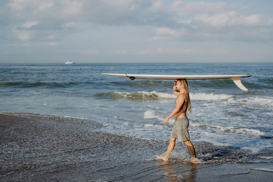 a young guy with dreadlocks carries a surfboard on his head, preparing to go riding the waves - Powered by Adobe