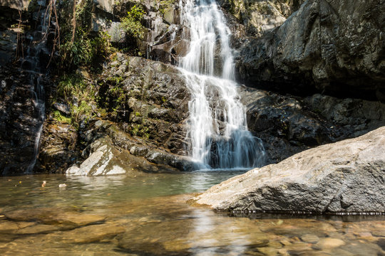 small waterfall run down  over the cliff wall under the sun with few green plants grown on the rocky surface