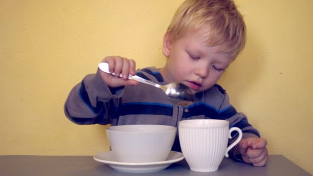 Small blond child sits at table with two cups on yellow background. Boy pours hot tea from one cup into another with spoon. Baby comes up with entertainment and is actively learning necessary skills