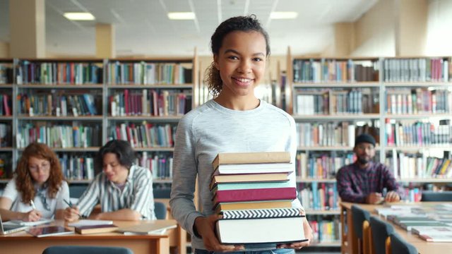 Happy African American woman student is walking in school library holding books smiling looking at camera. Education, youth and literature concept.