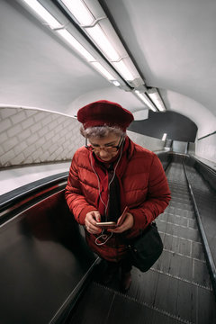 Senior woman standing at the escalator in Paris subway station, using smartphone with headphone