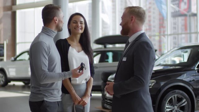 Happy beautiful wife and her cheerful husband speaking with salesman and taking key to their new car while buying in auto showroom