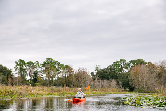 A pulled back image of senior man in his sixties paddling in the distance in an orange kayak on a marshy lake in Florida.