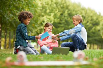 Fototapeta premium Two boys picking up the notebooks from girl while they sitting on the grass and doing homework together