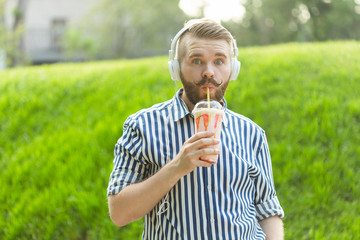 Fototapeta premium Side view of a stylish young man with a beard holding a milkshake and admiring the city views walking in the park on a warm summer day. The concept of rest and relaxation.