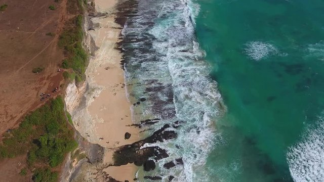 high up view of uluwatu cliffs and beach shot from drone, beautiful waves rolling over rocky beach, shot in bali, indonesia