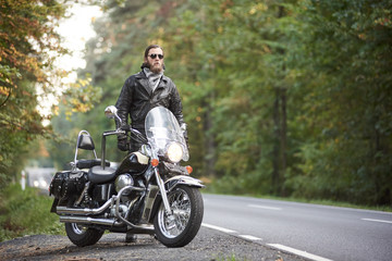 Fototapeta na wymiar Bearded tall motorcyclist in dark sunglasses, black leather jacket standing at shiny modern powerful cruiser motorbike on blurred background of hilly asphalt road and green trees.