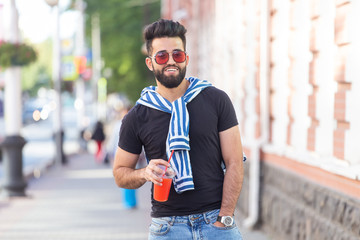 Handsome cheerful young arabian student with a stylish mustache and a beard with juice in his hands, walking around the city after a working day. Concept of positive and rest.