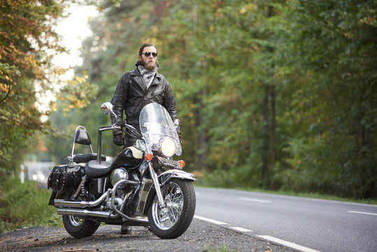 Bearded tall motorcyclist in dark sunglasses, black leather jacket standing at shiny modern powerful cruiser motorbike on blurred background of hilly asphalt road and green trees.