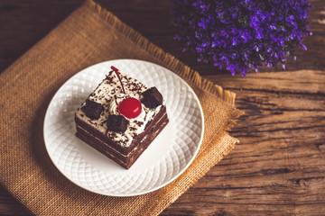 Black forest cake decorate with cherry and cake brownies In a white plate, place are on sackcloth and have false heather In pots small, all place are on wooden table.