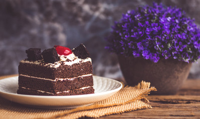 Black forest cake decorate with cherry and cake brownies In a white plate, place are on sackcloth and have false heather In pots small, all place are on wooden table.