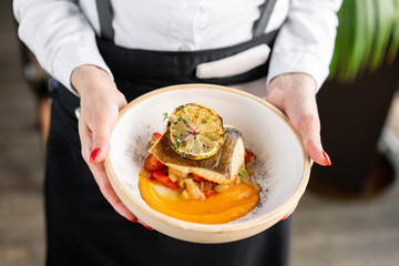 The waiter is holding a plate Roasted pike perch or cod fish with baked vegetables. Dish decorated with a slice of lemon. Restaurant menu