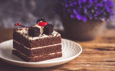 Black forest cake decorate with cherry and cake brownies In a white plate,and have false heather In pots small, all place are on wooden table.