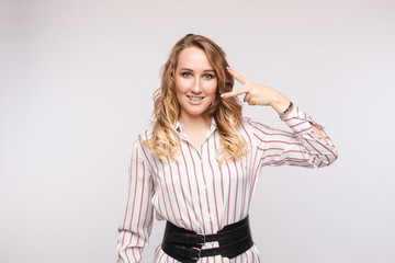 Studio portrait of pretty young Caucasian woman in striped shirt with fashionable wide waist belt showing peace sign near her face and smiling at camera. Isolate on white.