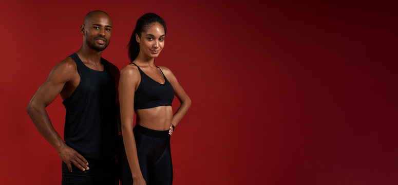 Training together. Young and cheerful african fitness couple in sportswear looking at camera with smile while standing against red background