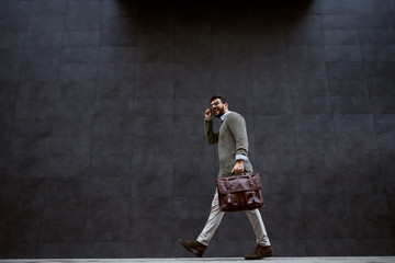 Full length of stylish caucasian designer in gray sweater holding leather bag while passing by gray...
