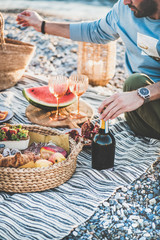 Summer beach picnic at sunset. Young couple sitting on blanket having weekend picnic outdoor at seaside with fresh seasonal fruit, tray of tasty appetizers and bottle of sparkling wine