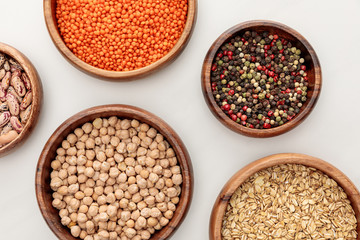 top view of wooden bowls with oatmeal, red lentil, peppercorns and chickpea on white marble surface