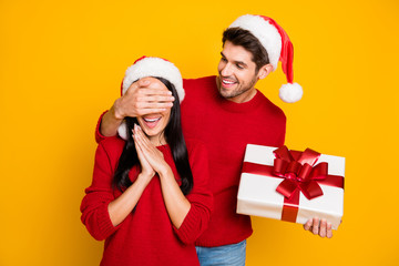 Portrait of handsome man with brown hair hide his excited wife eyes give her package wear red pullover isolated over yellow background