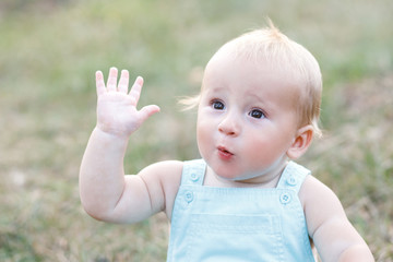 Cute blond boy sitting alone on the grass and waving his hand. The boy is ten months old