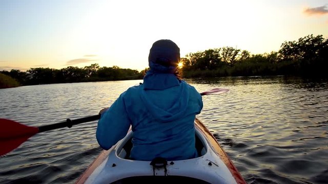 A woman is swimming in a kayak towards the setting sun on a calm river. She is rowing with oars. Slow motion