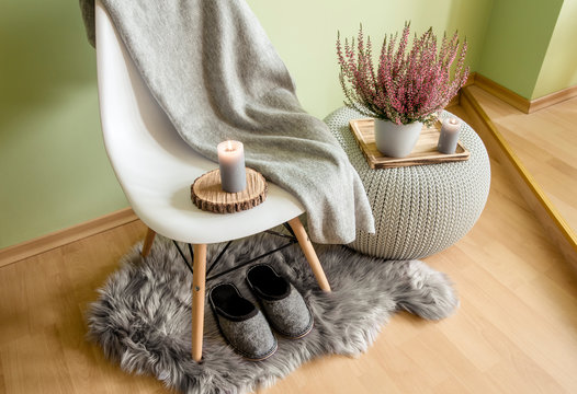 Plastic chair with wood legs covered with gray wool plaid, knitted pattern plastic table, fake sheepskin rug with felt slippers and heather in flower pot, against pale green wall.