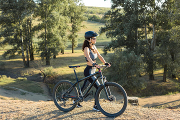 Fototapeta premium Girl on a mountain bike on offroad, beautiful portrait of a cyclist at sunset, Fitness girl rides a modern carbon fiber mountain bike in sportswear, a helmet, glasses and gloves. 