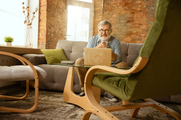 Senior man working with laptop at home - concept of home studying. Caucasian male model sitting on sofa and doing his homework or serfing in internet, watching cinema or webinar, playing games.