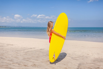 Back view of young woman with long hair in red swimwear holding the bright yellow surfboard on beach with sea and blue sky on background on Bali island, Indonesia. Active lifestyle. Holiday leisure.