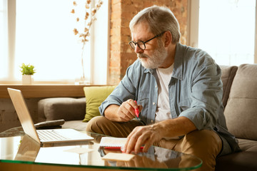 Senior man working with laptop at home - concept of home studying. Caucasian male model sitting on sofa and doing his homework or serfing in internet, watching cinema, making notes while webinar's on.