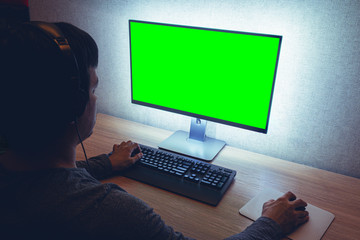 Man or gamer in headphones sits in front of a blank green chromakey monitor in dark room