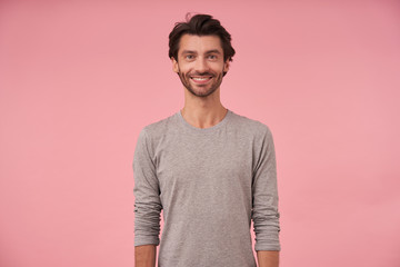 Studio shot of beautiful bearded male with dark hair standing over pink background, wearing casual clothes, looking at camera cheerfully with hands down