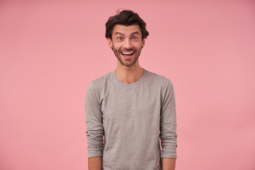 Indoor portrait of happy bearded male with trendy haircut wearing casual clothes, posing over pink background with hands down, looking at camera cheerfully with wide mouth opened
