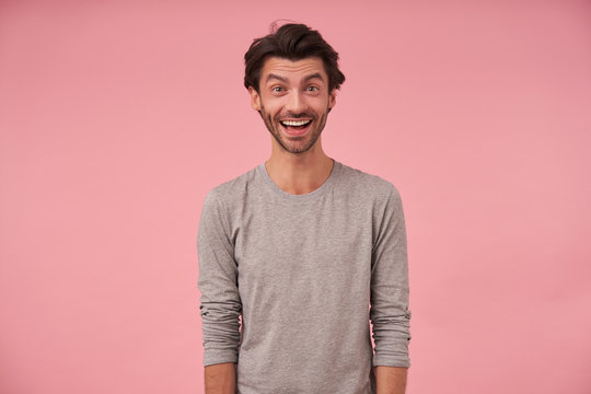 Indoor portrait of happy bearded male with trendy haircut wearing casual clothes, posing over pink background with hands down, looking at camera cheerfully with wide mouth opened