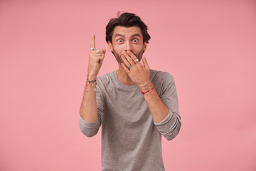 Surprised pretty bearded male with trendy haircut wearing grey sweater, standing over pink background with palm on his mouth, looking at camera and pointing upwards with index finger