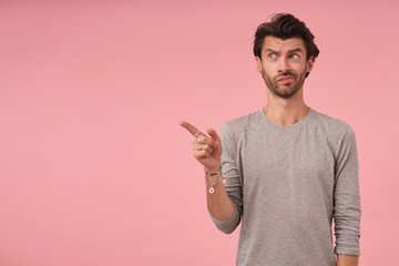 Studio photo of dark haired man with beard standing over pink background, pointing aside with forefinger, looking at camera with doubting face and pursing lips