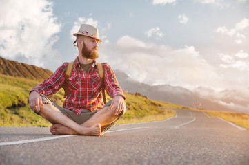 Stylish barefoot bearded male hitchhiker traveler in a hat and with a backpack sits on a suburban asphalt road in the mountains at sunset and waits for cash assistance looking to the sky.