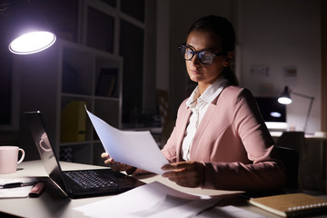 Serious manager in eyeglasses sitting at office desk and examining business contract she working till late evening at office