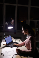 Young businesswoman sitting at the table using her laptop computer and writing on paper she working till late evening at office