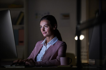 Young confident businesswoman sitting at the table in front of computer monitor and typing on...
